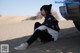 A woman sitting on the sand in front of a car.